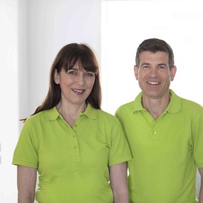 Zwei freundliche Mitarbeiter im grünen Polohemd stehen in heller Praxisumgebung.