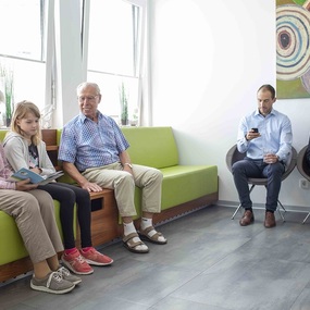 Personen unterschiedlichen Alters sitzen im Wartezimmer einer Arztpraxis und lesen.